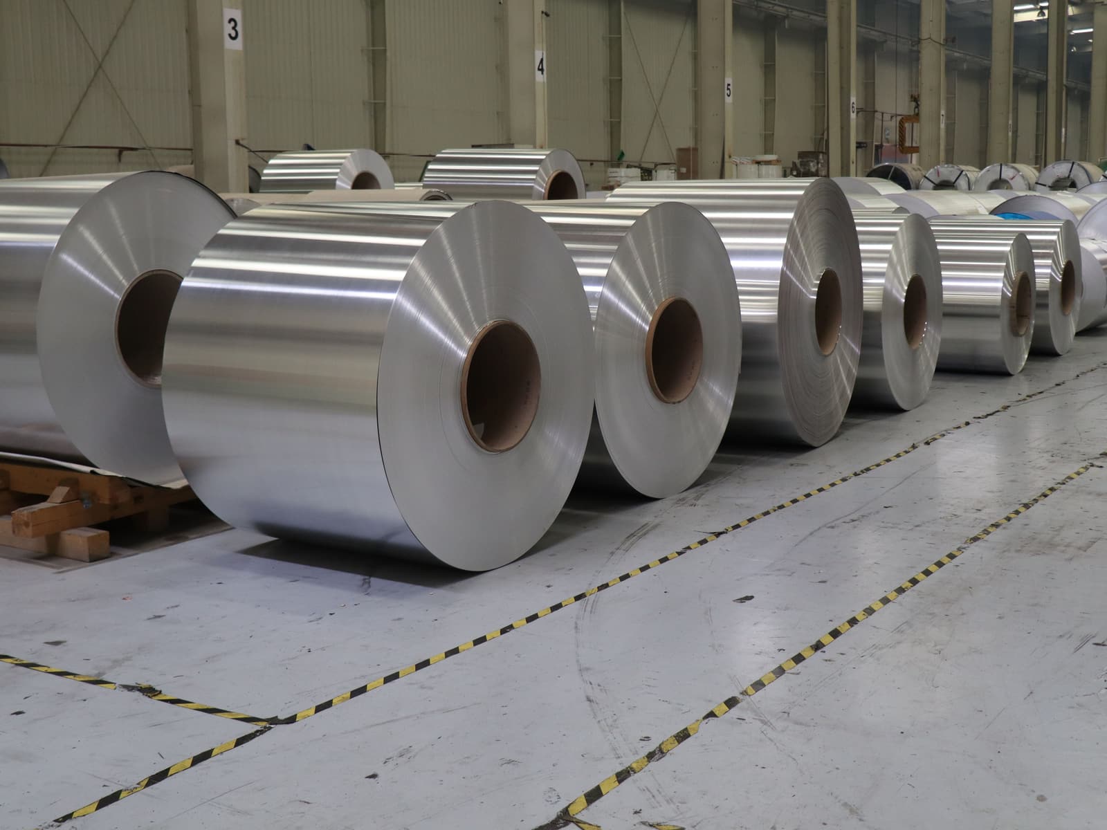 Fila di grandi bobine metalliche su un pavimento in cemento segnato da linee gialle e nere, in un capannone con colonne numerate