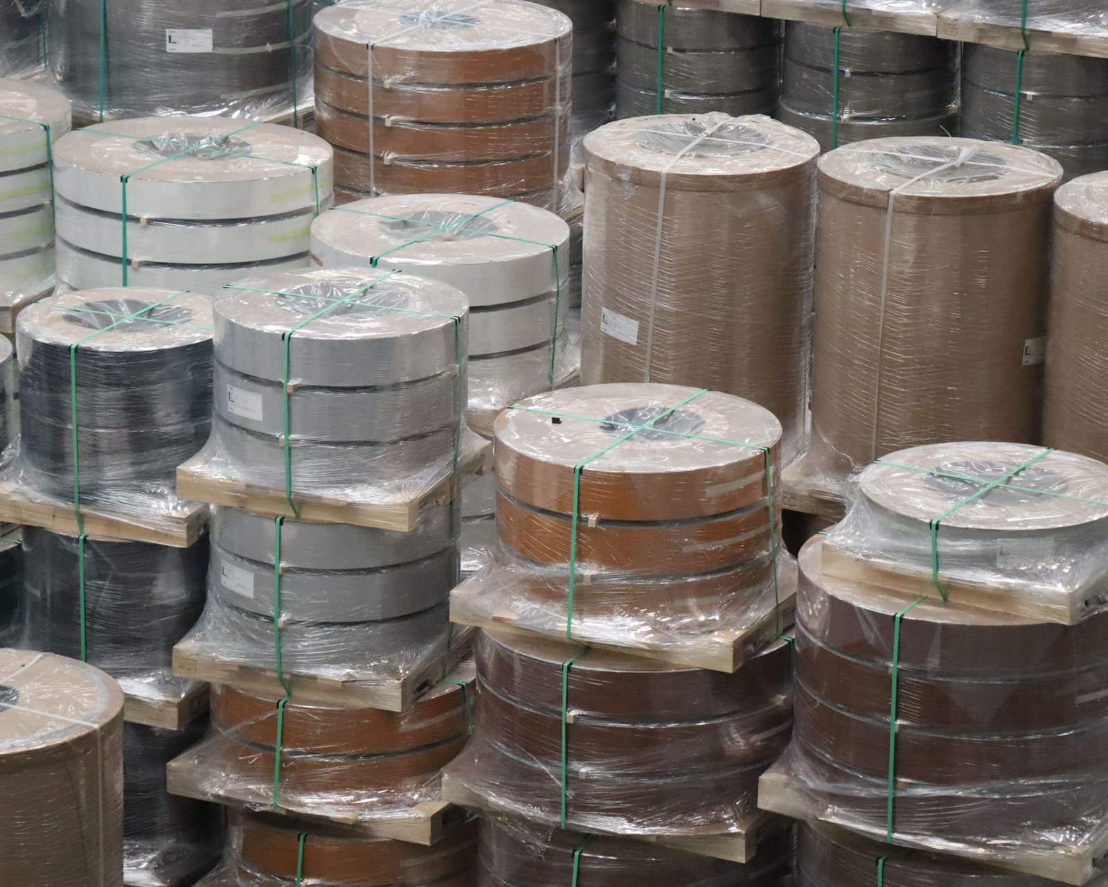 Pilhas de bobinas envolvidas em película transparente e presas com cintas verdes sobre paletes de madeira, com bordos em branco, cinzento, castanho e preto