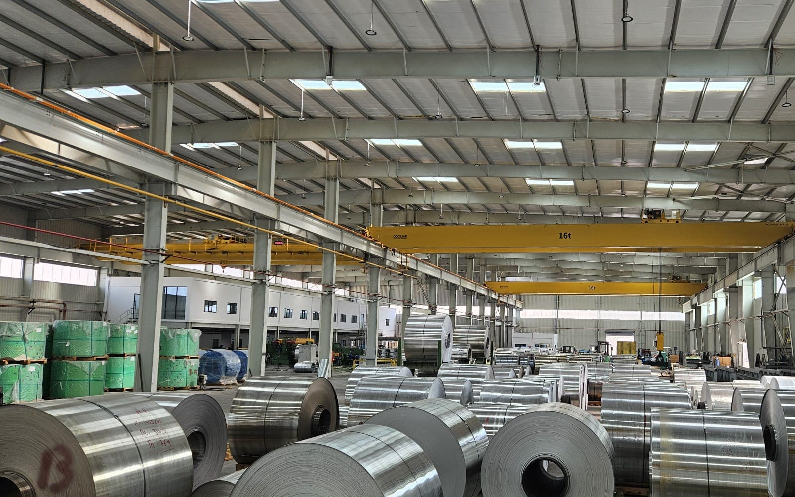 Large metal coils on wooden skids in a hall with yellow overhead cranes and green-wrapped stacks at the left