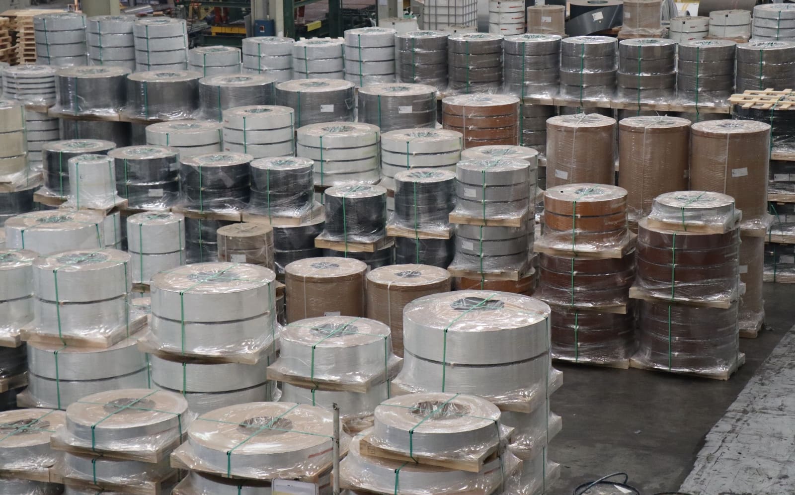 Rangées d'étroites bobines métalliques sur des palettes en bois, enveloppées de film plastique et cerclées, dans un entrepôt