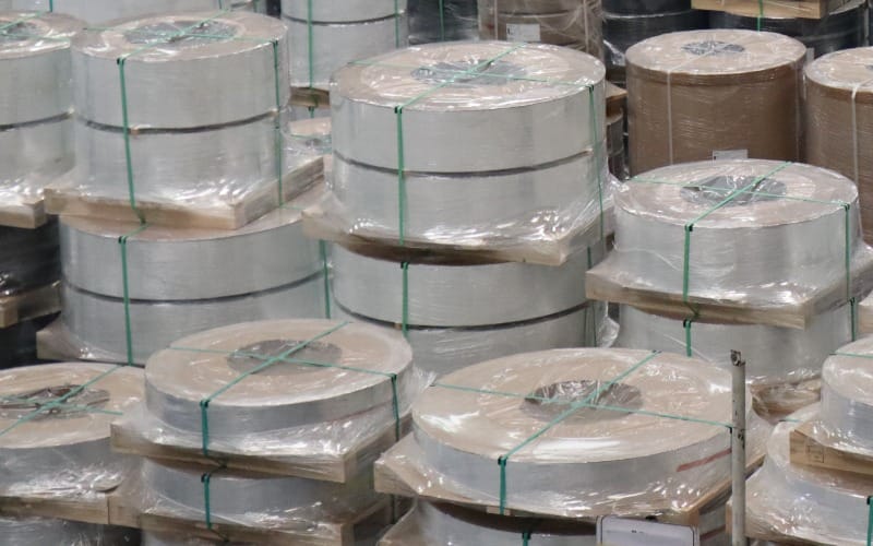 Narrow metal coils stacked on wooden pallets, wrapped in plastic film and held with green straps